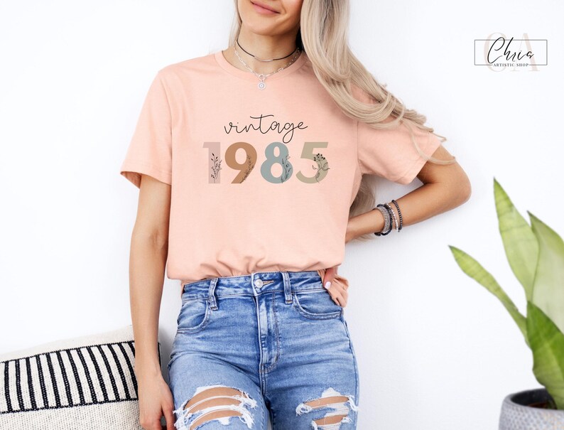 以下が含まれることがあります： 花柄のアクセントがついたスタイリッシュなフォントで「vintage 1985」と書かれた、ピーチ色のTシャツです。