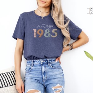 以下が含まれることがあります： 花柄のアクセントが付いたカラフルな文字で「ヴィンテージ 1985」と書かれたネイビーブルーのTシャツです。