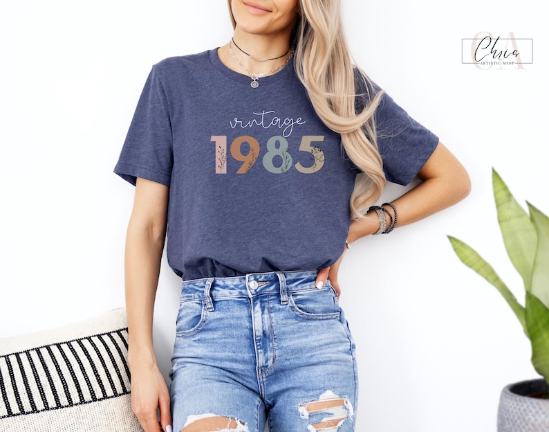 以下が含まれることがあります： 花柄のアクセントが付いたカラフルな文字で「ヴィンテージ 1985」と書かれたネイビーブルーのTシャツです。