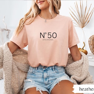 May include: Heather peach-colored t-shirt with the text "N&deg;50 1976 ORIGINAL" in black. The shirt is a short-sleeved crew neck. The model is wearing denim shorts and a cream-colored cardigan.