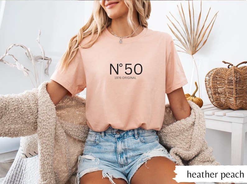May include: Heather peach-colored t-shirt with the text "N&deg;50 1976 ORIGINAL" in black. The shirt is a short-sleeved crew neck. The model is wearing denim shorts and a cream-colored cardigan.