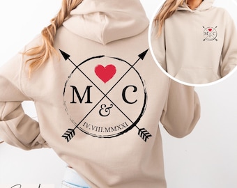 May include: A sand-colored hooded sweatshirt featuring a large graphic on the back and a smaller version on the front left chest. The design includes two crossed arrows, a red heart, the letters M and C, an ampersand, and the Roman numerals IV.VIII.MMXXI.