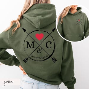 Könnte beinhalten: Olivgrüner Kapuzenpullover mit großem Aufdruck auf dem Rücken. Der Aufdruck zeigt ein rotes Herz, die Buchstaben "M" und "C" sowie die römische Zahl "IV.VIII.MMXXI". Ein kleineres, passendes Motiv befindet sich auf der Vorderseite. Das Wort "grün" steht unten links.