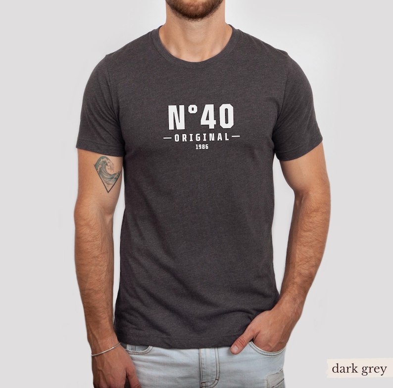 May include: A dark grey short-sleeved t-shirt featuring white text on the chest that reads N&deg;40 ORIGINAL 1986. The shirt has a crew neckline and a heathered fabric texture. The design is centered on the front of the garment.