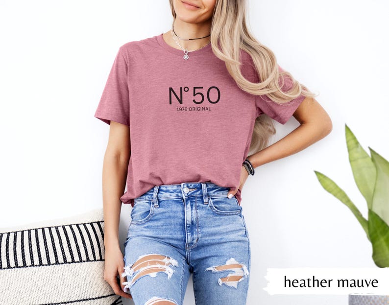 May include: Heather mauve t-shirt with the text "N&ordm; 50 1976 ORIGINAL" in black. The model is wearing blue jeans. The shirt is a solid color and has a round neckline. The shirt is made of a soft, comfortable material.