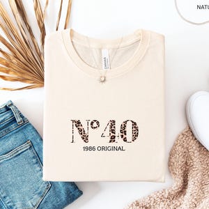 Könnte beinhalten: Cremefarbenes T-Shirt mit dem Text "N°40 1986 ORIGINAL" in Leopardenmuster. Das Shirt ist gefaltet und liegt auf einer weißen Oberfläche, neben einer blauen Jeans, weißen Turnschuhen und einem getrockneten Palmblatt.