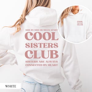 Könnte beinhalten: Weißer Kapuzenpullover mit dem Text "COOL SISTERS CLUB" in Staubrosa. Auf der Rückseite des Hoodies stehen die Sätze "SIDE BY SIDE OR MILES APART" und "SISTERS ARE ALWAYS CONNECTED BY HEART". Das Wort "WHITE" steht unten.