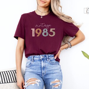 以下が含まれることがあります： ワインレッドのTシャツに、カラフルな花柄のフォントで「vintage 1985」と書いてあります。