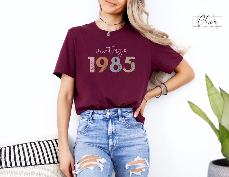 以下が含まれることがあります： ワインレッドのTシャツに、カラフルな花柄のフォントで「vintage 1985」と書いてあります。