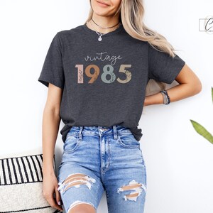 以下が含まれることがあります： 花柄のアクセントが付いたカラフルな文字で「ヴィンテージ 1985」と書かれたダークグレーのTシャツです。