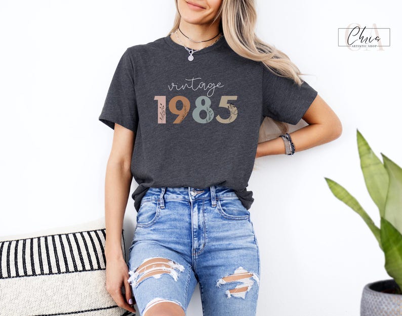 以下が含まれることがあります： 花柄のアクセントが付いたカラフルな文字で「ヴィンテージ 1985」と書かれたダークグレーのTシャツです。