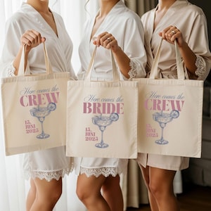 Könnte beinhalten: Drei cremefarbene Tragetaschen mit den Texten "Here comes the CREW" und "Here comes the BRIDE" in rosa Schrift. Jede Tasche zeigt eine Margarita-Illustration und das Datum "15. JUNI 2025" in Blau. Die Taschen werden von Personen gehalten.
