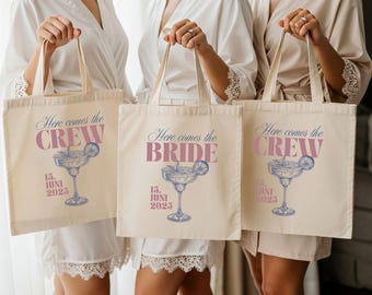 May include: Three cream-colored tote bags with the text "Here comes the CREW" and "Here comes the BRIDE" in pink script. Each bag features a margarita illustration and the date "15. JUNI 2025" in blue. The bags are held by people.