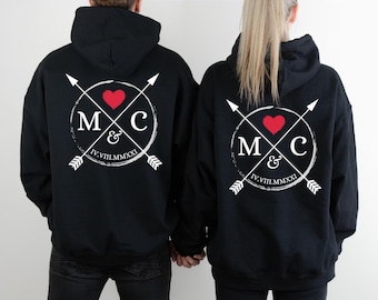 May include: Two black hooded sweatshirts with a white circle design on the back. The circle contains the letters "M & C" with a red heart above the letters and two white arrows crossing through the circle. The text "IV.VIII.MMXXI" is below the letters.