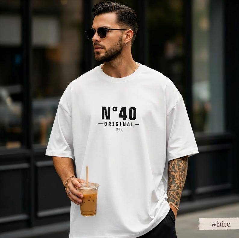 May include: A white short-sleeved t-shirt featuring black text on the chest that reads N&deg;40, ORIGINAL, and 1986. The shirt has a crew neckline and a loose, relaxed fit. The design is centered on the front of the garment in a bold, sans-serif font.