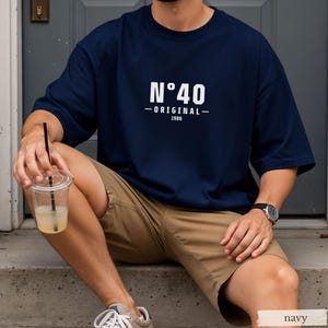 May include: A navy blue short-sleeved t-shirt featuring white printed text that reads N&deg;40 ORIGINAL 1986 centered on the chest. The shirt has a crew neckline and a loose fit, paired with khaki shorts, light-colored sneakers, and a wristwatch.