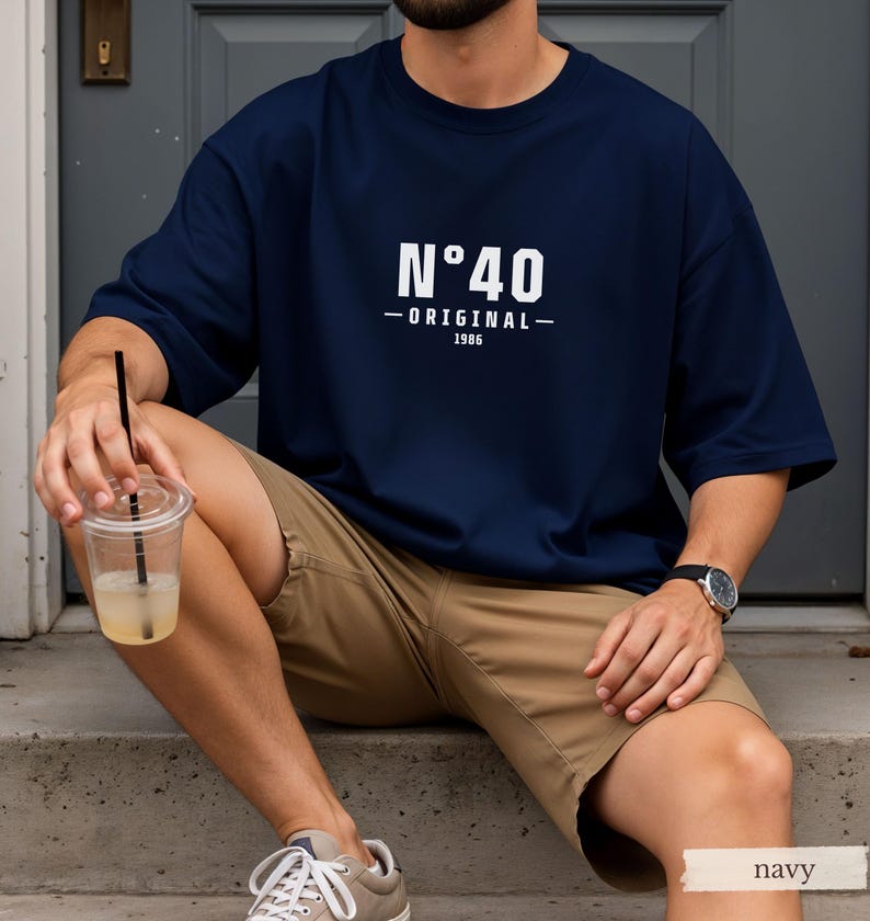 May include: A navy blue short-sleeved t-shirt featuring white printed text that reads N&deg;40 ORIGINAL 1986 centered on the chest. The shirt has a crew neckline and a loose fit, paired with khaki shorts, light-colored sneakers, and a wristwatch.