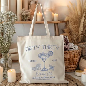 May include: Cream-colored tote bag with blue text and a cocktail illustration. The text reads "Dirty Thirty," "October 1995," and "Birthday Club Aged to Perfection." The bag has long handles.