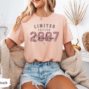 K&ouml;nnte beinhalten: Heather-Peachfarbenes T-Shirt mit dem Text "LIMITED EDITION 2007 Carolina" in Grau und Schwarz. Das Shirt hat eine kleine Herzgrafik. Das Model tr&auml;gt Jeansshorts und eine beige Strickjacke.
