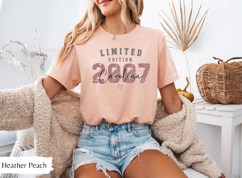 K&ouml;nnte beinhalten: Heather-Peachfarbenes T-Shirt mit dem Text "LIMITED EDITION 2007 Carolina" in Grau und Schwarz. Das Shirt hat eine kleine Herzgrafik. Das Model tr&auml;gt Jeansshorts und eine beige Strickjacke.