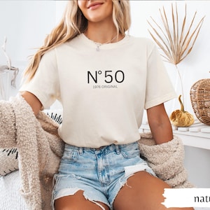 May include: Cream-colored t-shirt with the number "50" and the text "1976 ORIGINAL" in black. The model is wearing denim shorts and a beige cardigan. The word "natural" is visible in the bottom right corner.