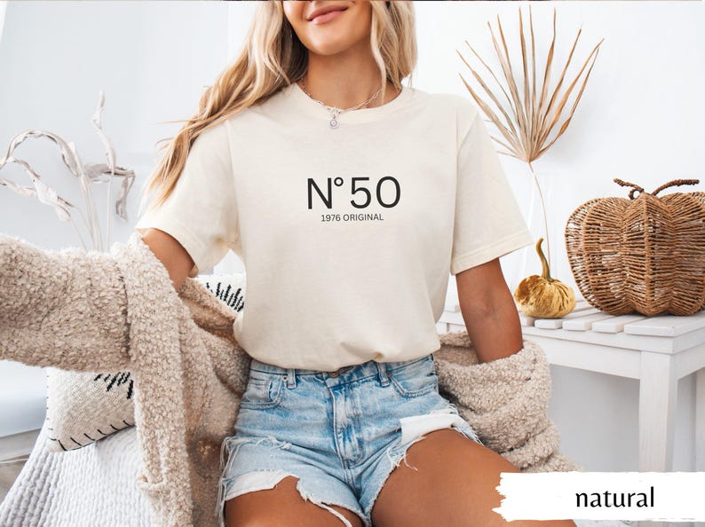 May include: Cream-colored t-shirt with the number "50" and the text "1976 ORIGINAL" in black. The model is wearing denim shorts and a beige cardigan. The word "natural" is visible in the bottom right corner.