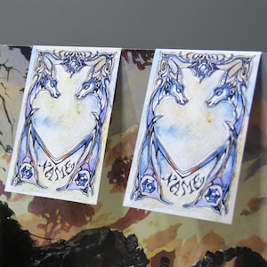 Puede incluir: Tres ilustraciones de acuarela de un marco en forma de corazón con un par de dragones estilizados a cada lado. Los dragones son azules, morados y blancos. El texto "Nombre" está escrito en una fuente decorativa en la parte inferior de cada marco.