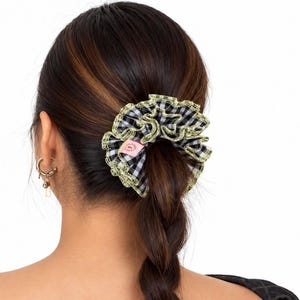 May include: A black and white gingham patterned hair scrunchie featuring a ruffled edge trimmed with a light green lace border. A small pink rectangular fabric tag is attached to the inner side of the scrunchie, securing a section of hair.