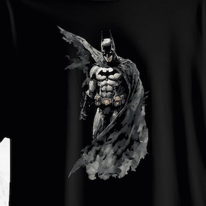 May include: A black t-shirt featuring a graphic illustration of Batman in a dark, painterly style. The character is depicted in a black and grey suit with a flowing cape, a utility belt, and the iconic bat emblem centred on the chest area.