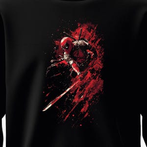 Pode incluir: Camisola preta com um gráfico dinâmico de um super-herói em um traje vermelho e preto, empunhando uma espada. O design incorpora efeitos de respingos vermelhos, criando uma sensação de movimento. O super-herói está em posição de agachamento, pronto para a batalha.