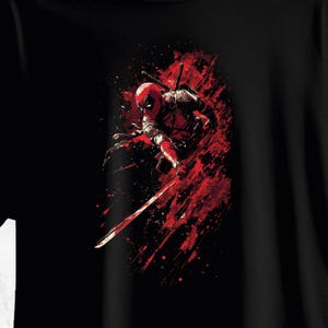 Pode incluir: T-shirt preta com uma ilustração dinâmica de Deadpool em vermelho e preto. O personagem é retratado em uma pose de luta com uma espada, contra um fundo de respingos vermelhos.