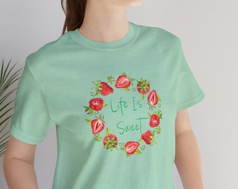 Op de afbeelding: Een lichtgroen t-shirt met een afbeelding van aardbeien in een cirkel rond de tekst "Life Is Sweet".
