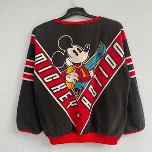 May include: Black sweatshirt featuring Mickey Mouse holding a football, with the words "MICKEY ACTION" in a red and white chevron design. The sweatshirt has red trim and sleeve stripes.