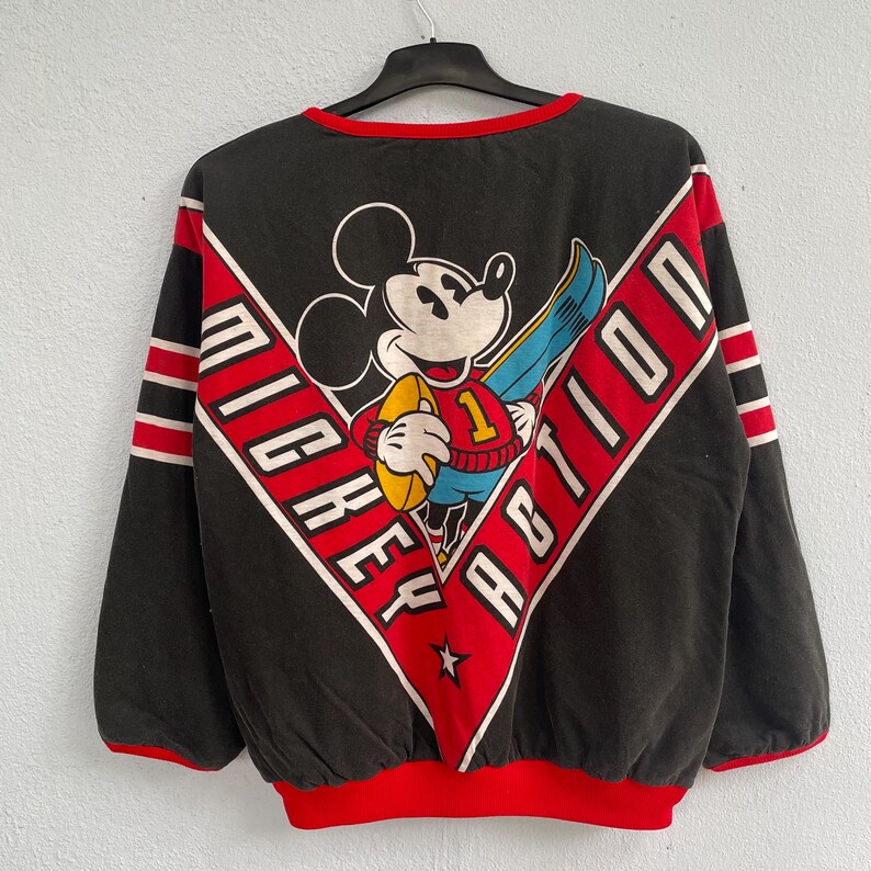 May include: Black sweatshirt featuring Mickey Mouse holding a football, with the words "MICKEY ACTION" in a red and white chevron design. The sweatshirt has red trim and sleeve stripes.