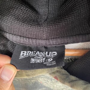 May include: Close-up of a black tag with the words "BREAKUP" and "INFINITY" in white. Below the text is "MOSSY OAK BRAND CAMO". The tag is attached to a camouflage hooded sweatshirt.
