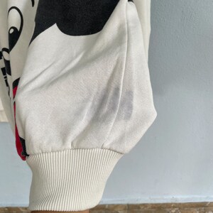 Puede incluir: Una sudadera de manga larga en color blanco roto con un gran estampado gr&aacute;fico negro en el lateral y un detalle en rojo. La prenda tiene un pu&ntilde;o acanalado en la mu&ntilde;eca y presenta una tenue mancha gris en la tela cerca de la manga.