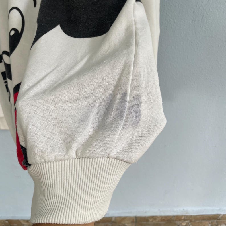 Puede incluir: Una sudadera de manga larga en color blanco roto con un gran estampado gr&aacute;fico negro en el lateral y un detalle en rojo. La prenda tiene un pu&ntilde;o acanalado en la mu&ntilde;eca y presenta una tenue mancha gris en la tela cerca de la manga.