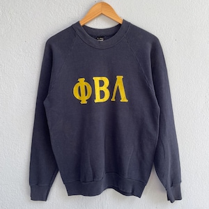 Peut inclure: Sweat-shirt bleu marine avec les lettres grecques ΦΒΛ en jaune. Le sweat-shirt à col rond est suspendu à un cintre en bois. Les lettres sont grandes et centrées sur le devant du vêtement.