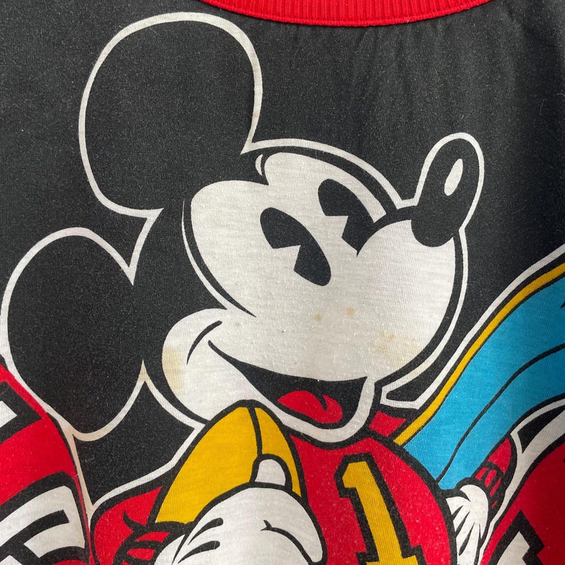 May include: Black t-shirt featuring a large graphic of Mickey Mouse. The design includes Mickey's face in white, outlined in black, with a red mouth. The shirt has a red collar and accents of yellow and blue.