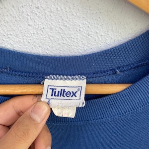 Peut inclure: Un sweat-shirt bleu &agrave; col rond avec une &eacute;tiquette en tissu blanc au niveau du col int&eacute;rieur. L'&eacute;tiquette affiche le nom de la marque Tultex en bleu avec un symbole de marque d&eacute;pos&eacute;e. Le v&ecirc;tement est fait d'un tissu bleu uni avec un col c&ocirc;tel&eacute;.
