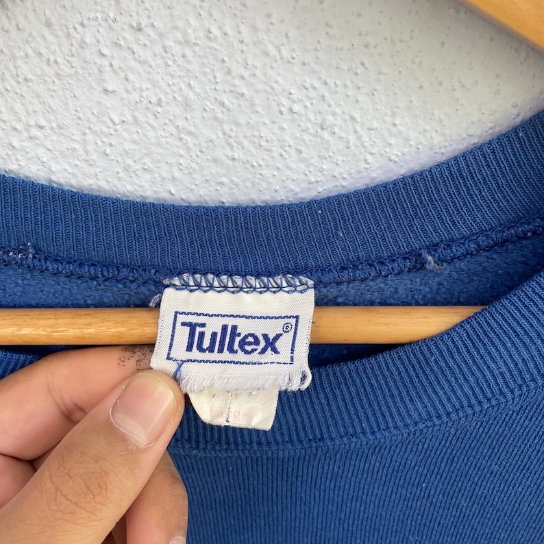 Peut inclure: Un sweat-shirt bleu &agrave; col rond avec une &eacute;tiquette en tissu blanc au niveau du col int&eacute;rieur. L'&eacute;tiquette affiche le nom de la marque Tultex en bleu avec un symbole de marque d&eacute;pos&eacute;e. Le v&ecirc;tement est fait d'un tissu bleu uni avec un col c&ocirc;tel&eacute;.