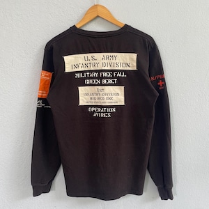 May include: A black long-sleeve shirt featuring white rectangular patches with black text reading U.S. ARMY INFANTRY DIVISION, MILITARY FREE FALL, GREEN BERET, 1ST INFANTRY DIVISION BIG RED ONE, and OPERATION AVIREX. An orange patch is on the left sleeve.