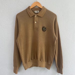 May include: A tan long-sleeve polo shirt featuring a ribbed knit texture, a collar with two buttons, and a black circular patch on the chest with the text Lacoste Club and a green crocodile logo. The shirt includes a white interior neck label.