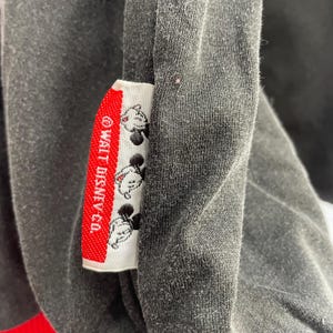May include: Close-up of a dark gray garment with a red and white tag. The tag features the text "&copy; WALT DISNEY CO." and several Mickey Mouse illustrations. The garment appears to be made of a soft, textured fabric.