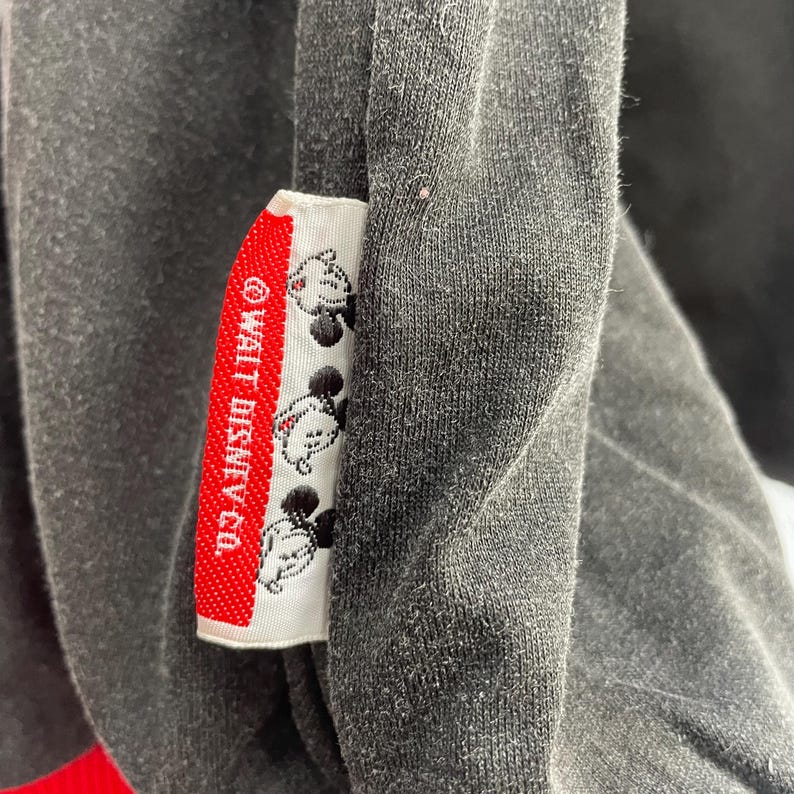 May include: Close-up of a dark gray garment with a red and white tag. The tag features the text "&copy; WALT DISNEY CO." and several Mickey Mouse illustrations. The garment appears to be made of a soft, textured fabric.