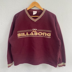 May include: A maroon long-sleeve sweatshirt with a V-neck collar featuring tan and dark brown stripes. The chest displays the Billabong logo in tan lettering with the text "since nineteen seventy three" underneath, all enclosed by horizontal lines.