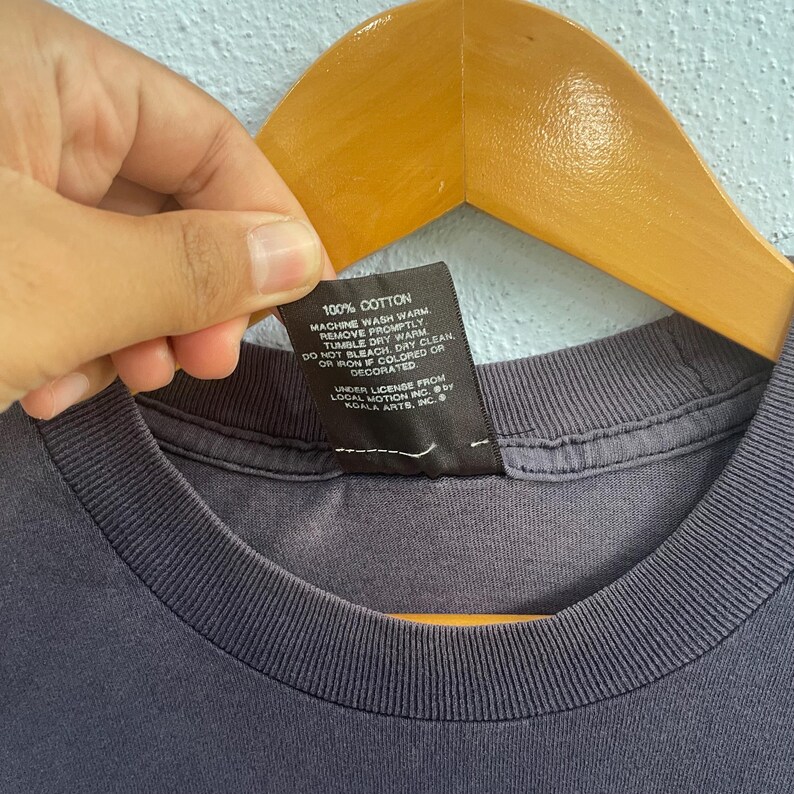Op de afbeelding: Een zwart verzorgingsetiket met witte tekst die luidt: "100% Katoen Machinewas warm Onmiddellijk verwijderen Droogtrommel warm Niet bleken Stomen toegestaan Niet strijken als gekleurd of versierd Licentie van Local Motion Inc. door Koala Arts Inc."