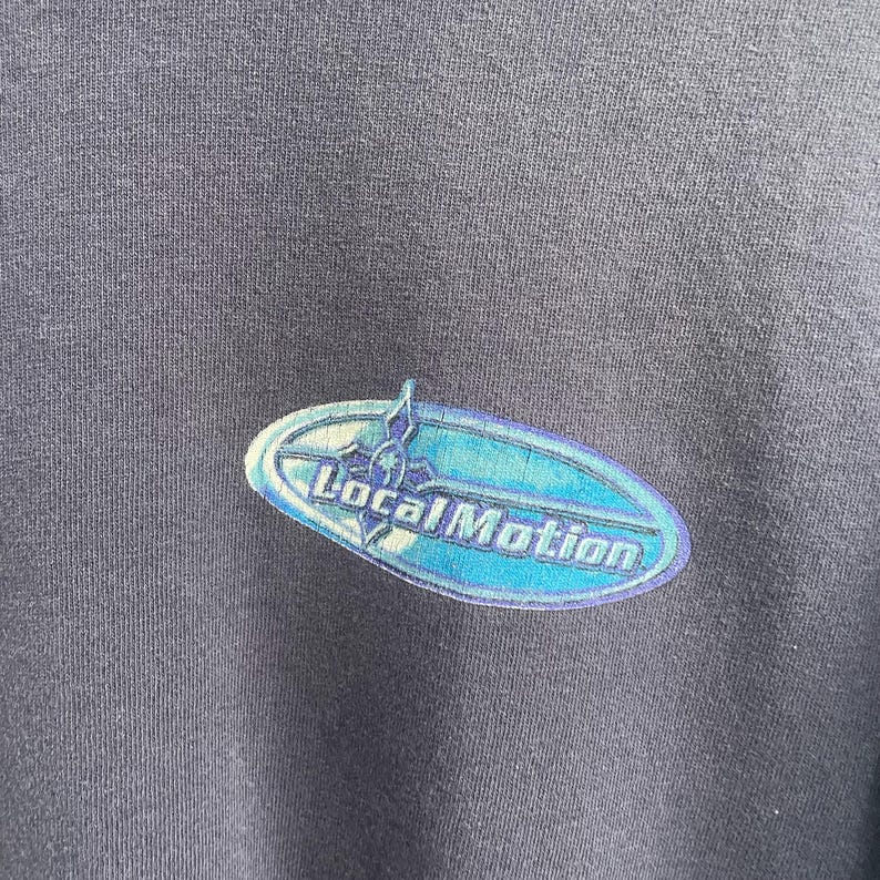 Op de afbeelding: Een donkergrijs T-shirt met een blauwe en witte afbeelding van een surfboard met de tekst "Local Motion" erop.