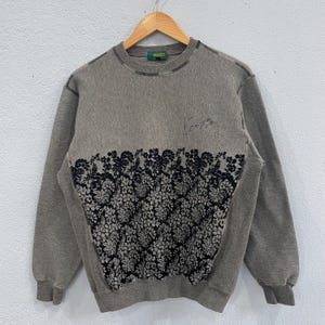 May include: A gray sweatshirt with a floral design across the chest. The floral pattern is black and white. The word "Kenzo" is on the tag and the name "Kenzo" is written in cursive on the upper chest.