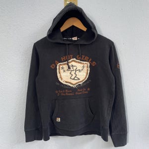 Op de afbeelding: Zwarte hoodie met een kangoeroezak en trekkoord. De voorkant heeft een afbeelding met de tekst "DA HUT GIRLS SINCE 1975" en andere tekst. De hoodie heeft een capuchon en lange mouwen.